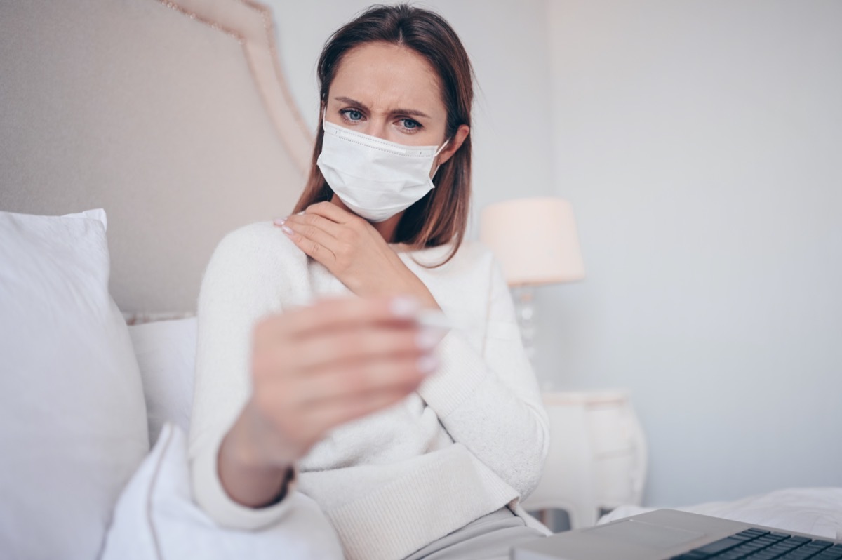 Sick woman in face protection mask lying in bed holding thermometer at home quarantine isolation