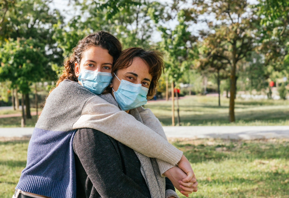 A daughter hugs her mother from behind in a park as both wear face masks
