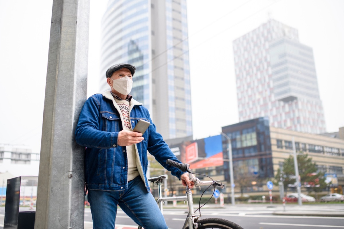 older man with face mask and phone standing next to his bike outside